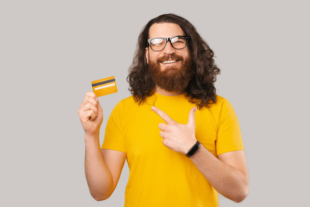 Joyful bearded man wearing glasses and pointing at yellow debit card over grey backdrop - Zdjęcie, obraz