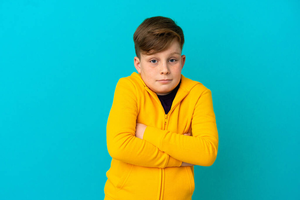 Kleine roodharige jongen geïsoleerd op blauwe achtergrond het maken van twijfels gebaar terwijl het optillen van de schouders - Foto, afbeelding
