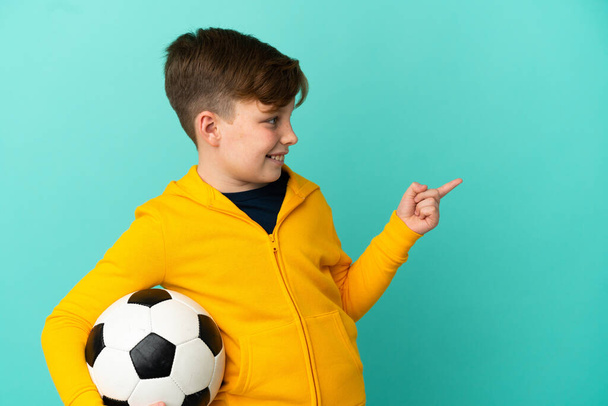 Punapää lapsi pelaa jalkapalloa eristetty sinisellä taustalla osoittaa sormella sivulle ja esittää tuotteen - Valokuva, kuva
