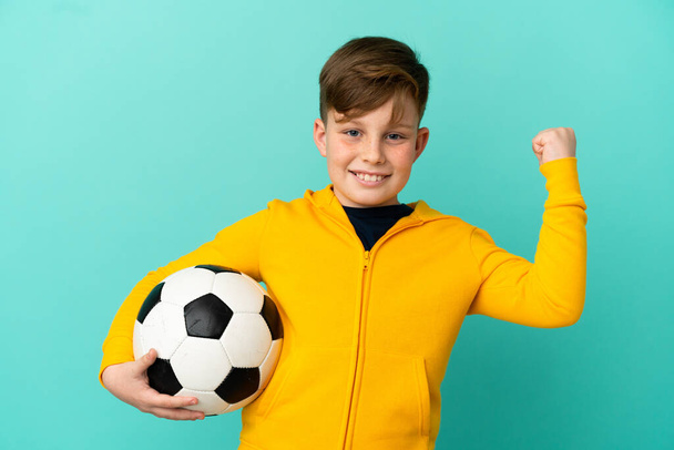 Redhead παιδί παίζει ποδόσφαιρο απομονωμένο σε μπλε φόντο γιορτάζει μια νίκη - Φωτογραφία, εικόνα