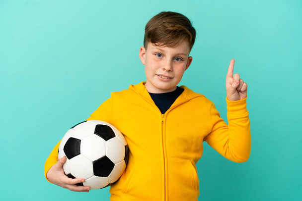 Pelirroja jugando fútbol aislado sobre fondo azul mostrando y levantando un dedo en señal de la mejor - Foto, imagen
