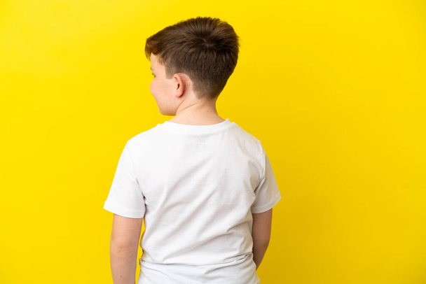 Little redhead boy isolated on yellow background in back position and looking side - Photo, image