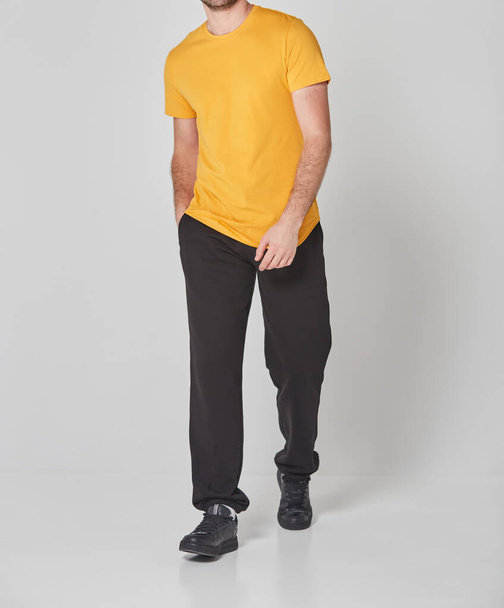 Foto recortada de hombre en camiseta en blanco amarillo y pantalones negros. De pie sobre fondo gris. Mockup para imprimir o plantilla de diseño. Línea de ropa básica sin logotipo - Foto, Imagen