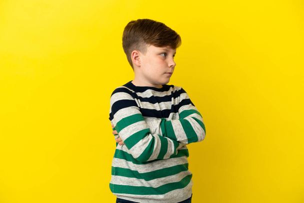 Μικρό κοκκινομάλλικο αγόρι απομονωμένο σε κίτρινο φόντο κρατώντας σταυρωμένα τα χέρια - Φωτογραφία, εικόνα