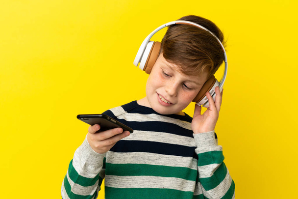 Little redhead boy isolated on yellow background listening music with a mobile and singing - Photo, Image