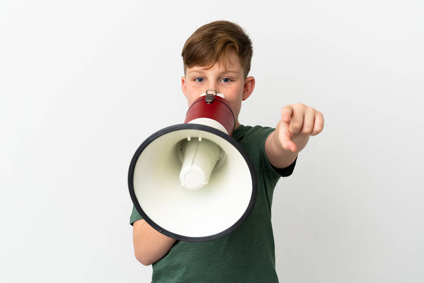Μικρό κοκκινομάλλικο αγόρι απομονωμένο σε λευκό φόντο φωνάζοντας μέσα από ένα μεγάφωνο για να ανακοινώσει κάτι ενώ δείχνει προς τα εμπρός - Φωτογραφία, εικόνα