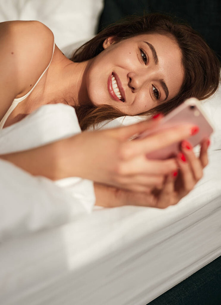 Delighted young woman using smartphone while lying in bed in morning - Φωτογραφία, εικόνα