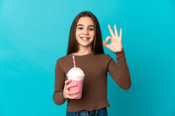 Little girl with strawberry milkshake isolated on blue background showing ok sign with fingers - Fotó, kép