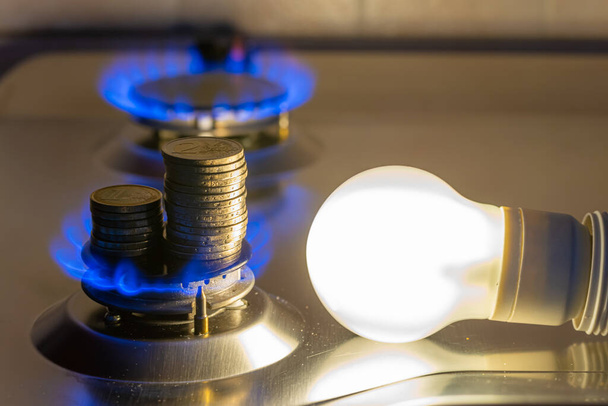 Vedle zapáleného plynového sporáku se rozsvítí žárovka a vedle ní mince. Náklady na energii a plyn, zvýšení nákladů. - Fotografie, Obrázek