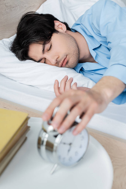 Человек с закрытыми глазами лежит в постели и выключает размытый будильник - Фото, изображение