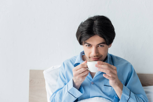 brunette man glimlachen op camera tijdens het drinken van koffie in de slaapkamer - Foto, afbeelding