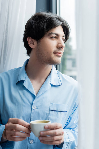 young brunette man in blue pajamas standing with coffee cup near window - Zdjęcie, obraz