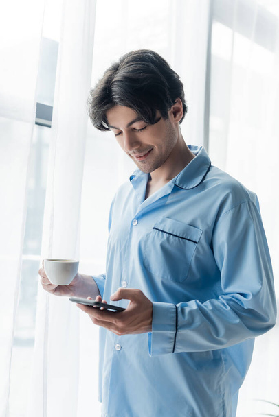 positive man in blue pajamas holding coffee cup and chatting on smartphone near window - Foto, Imagen