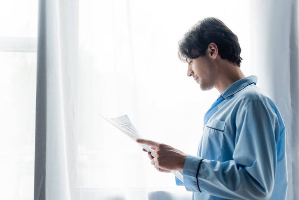 side view of young smiling man in blue pajamas reading morning newspaper near window - Photo, Image