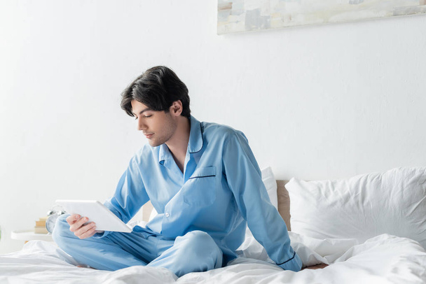 young man in blue pajamas looking at digital tablet while sitting on bed with crossed legs - Fotoğraf, Görsel