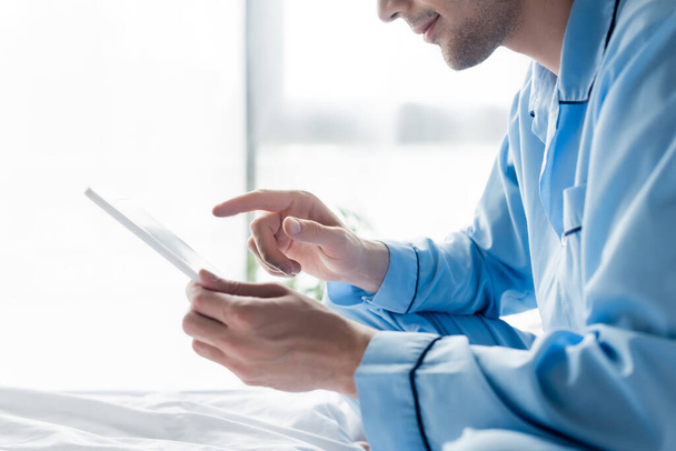 vista parcial del hombre en pijama azul usando tableta digital en el dormitorio - Foto, imagen
