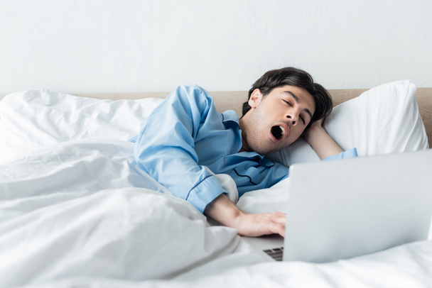 sleepy man yawning near laptop while lying in bed in morning - Foto, afbeelding