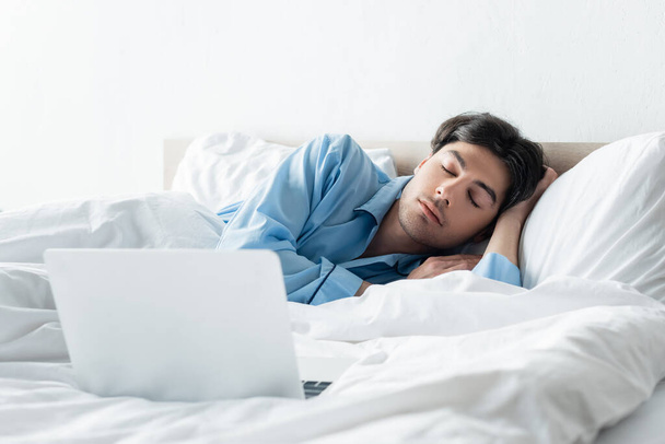 brunette man in blue pajamas sleeping near laptop in morning - Foto, afbeelding