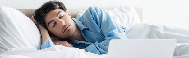 brunette man in blue pajamas sleeping on white bedding near laptop, banner - Photo, Image