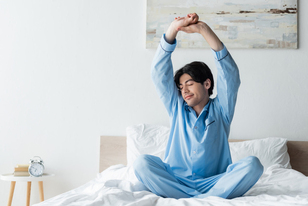 glimlachende man in blauwe pyjama die armen uitstrekt op bed terwijl hij met gekruiste benen zit - Foto, afbeelding
