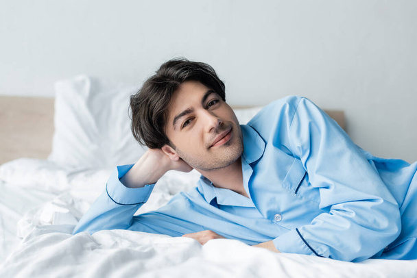 pleased man in blue pajamas looking at camera while lying on white bedding - Foto, afbeelding