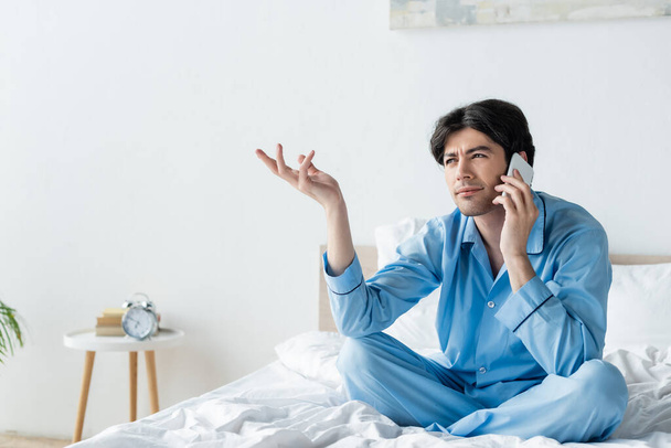 thoughtful man sitting on bed with crossed legs and gesturing during conversation on smartphone - Foto, Imagem