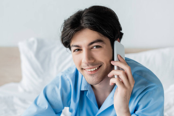 pleased man looking at camera while talking on mobile phone in bedroom - Fotó, kép