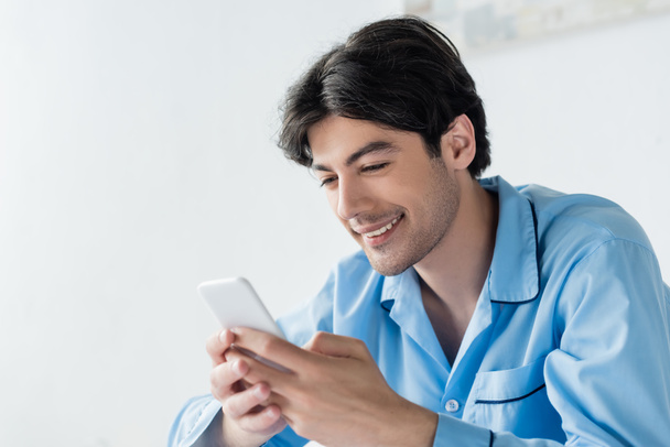 happy brunette man in blue pajamas chatting on mobile phone - Foto, Imagen