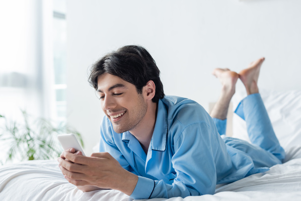 cheerful barefoot man in blue pajamas messaging on mobile phone in bedroom - Фото, изображение