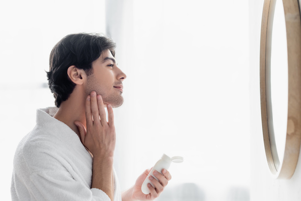 side view of brunette man applying cosmetic milk on face in bathroom - Photo, Image
