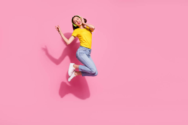 Kuva makea unenomainen nuori nainen käyttää keltainen asu hyppäämällä korkea kuuntelee kappaleita kuulokkeet tyhjä tila eristetty vaaleanpunainen väri tausta - Valokuva, kuva