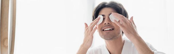 smiling brunette man wiping eyes with cotton pads in bathroom, banner - Foto, afbeelding