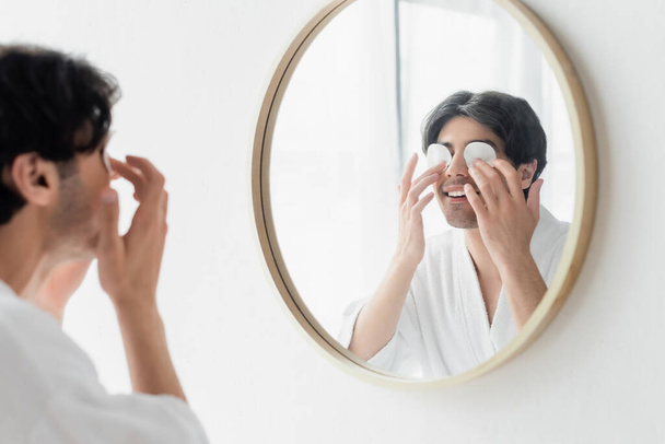hombre sonriente en albornoz blanco limpiando los ojos cerca del espejo en el baño - Foto, Imagen