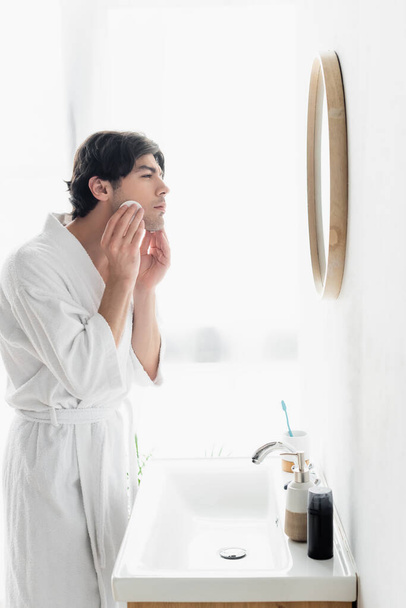 vista lateral del hombre en la cara de limpieza de albornoz blanco con almohadilla de algodón en el baño - Foto, imagen
