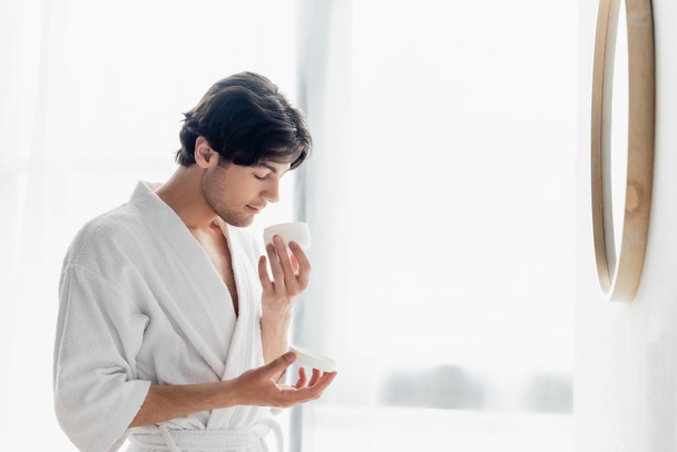 young man in bathrobe smelling cosmetic cream in bathroom - Photo, Image
