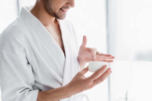 cropped view of man in white bathrobe holding container with cosmetic cream - Photo, Image