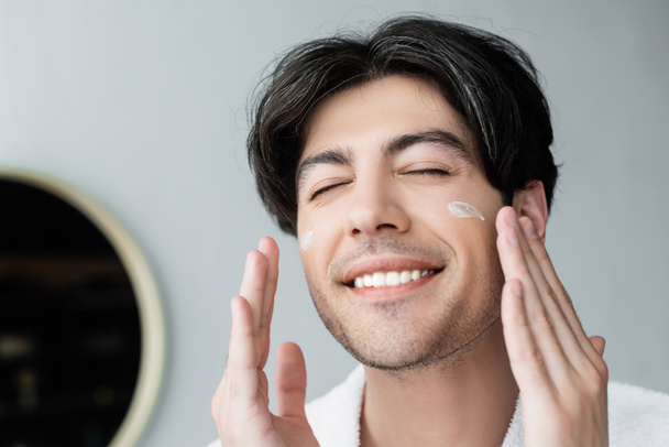 happy man with closed eyes applying face cream in bathroom - Photo, Image