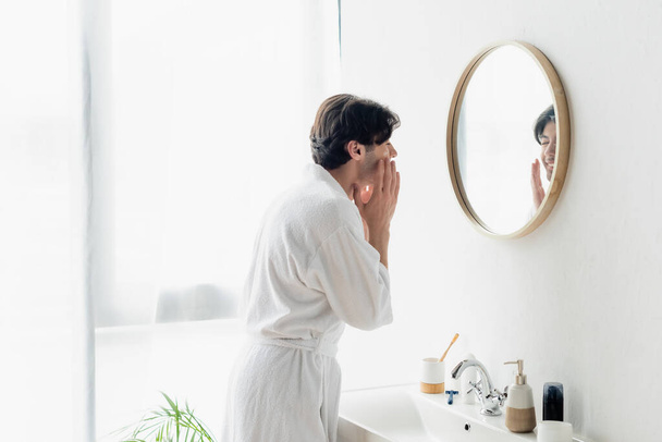 brunette homme en peignoir blanc appliquant crème visage près du miroir et de l'évier - Photo, image