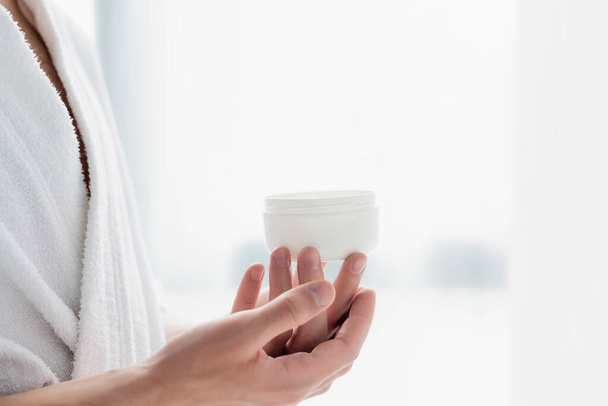 cropped view of man in bathrobe holding container with cosmetic cream - Foto, imagen