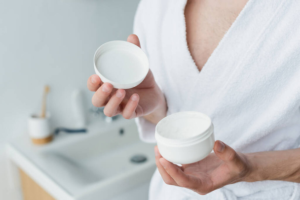 cropped view of man in white bathrobe holding cosmetic cream in blurred bathroom - Foto, Bild
