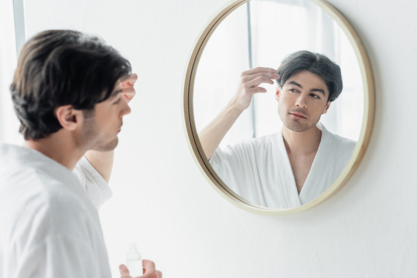 young man in white bathrobe applying cosmetic serum near mirror in bathroom - Photo, Image
