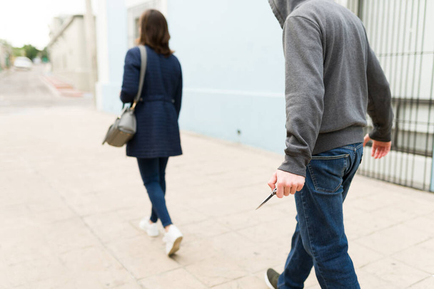 Vista trasera de un criminal usando un cuchillo para amenazar y robar a una joven caminando sola en la calle - Foto, Imagen