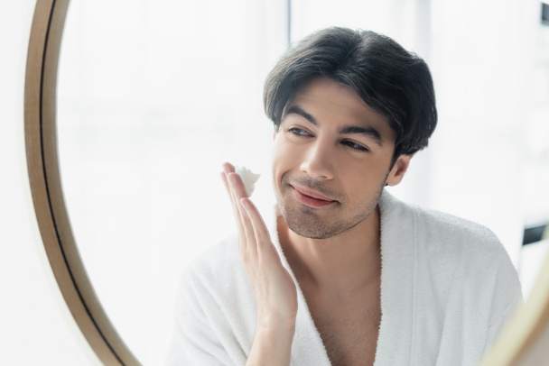 joven hombre sonriente en albornoz aplicando espuma de afeitar cerca del espejo - Foto, Imagen