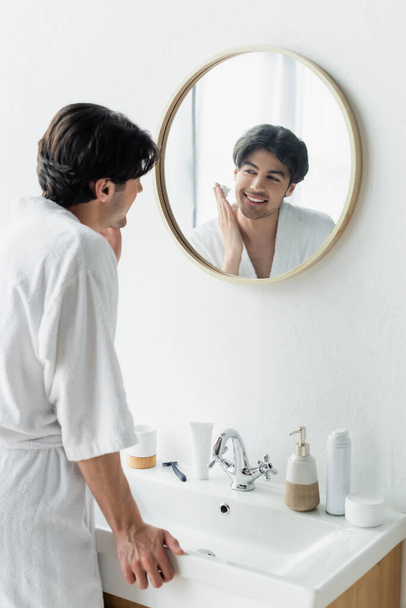 uomo sorridente che applica schiuma da barba vicino a specchio, articoli da toeletta e rasoio di sicurezza sul lavandino - Foto, immagini