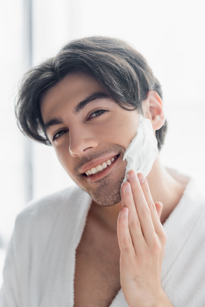 cheerful brunette man smiling at camera while applying shaving foam - Zdjęcie, obraz