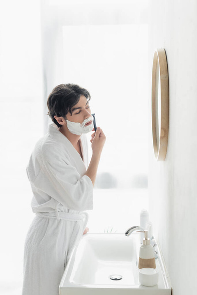 hombre joven en albornoz blanco afeitándose cerca del espejo y fregadero en el baño - Foto, imagen