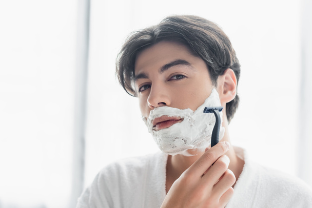 brunette man looking at camera while shaving with safety razor - Photo, Image
