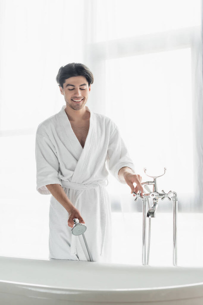 homem sorridente em roupão de banho branco segurando cabeça de chuveiro ao abrir a torneira no banheiro - Foto, Imagem