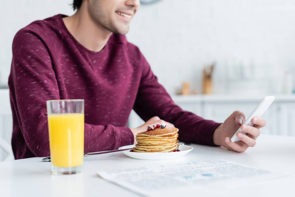 bijgesneden weergave van glimlachende man met smartphone in de buurt van pannenkoeken en sinaasappelsap - Foto, afbeelding