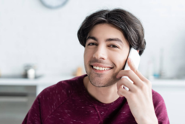happy brunette man talking on mobile phone and looking at camera at home - Foto, imagen